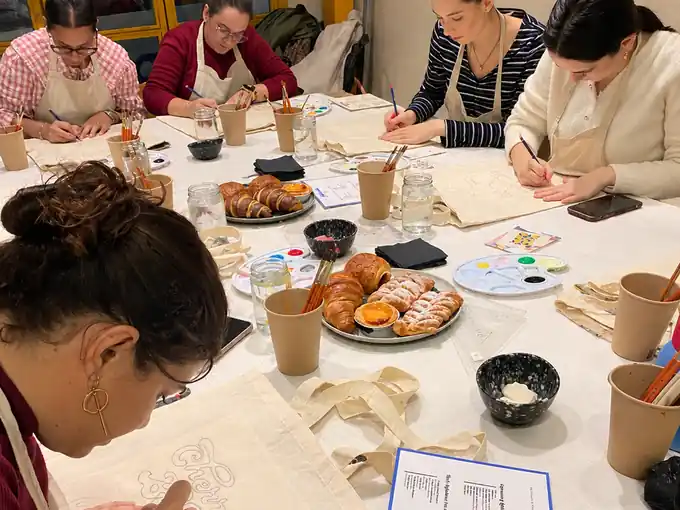 Participantes pintando tote bags con dulces y café en la mesa